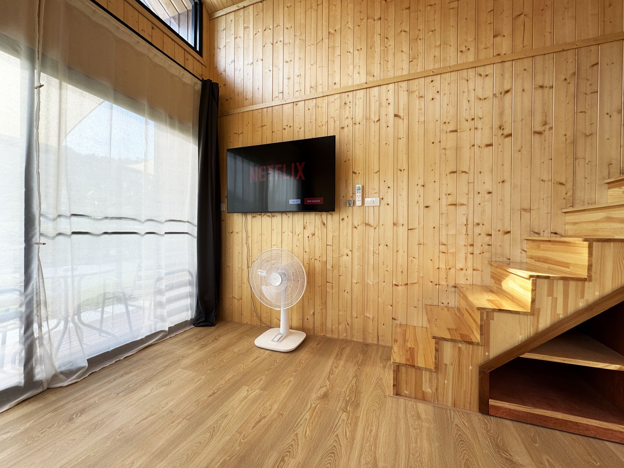 A room with wooden panel walls, a flat-screen TV mounted on the wall, a standing fan, and a staircase.