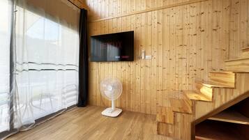 A room with wooden panel walls, a flat-screen TV mounted on the wall, a standing fan, and a staircase.