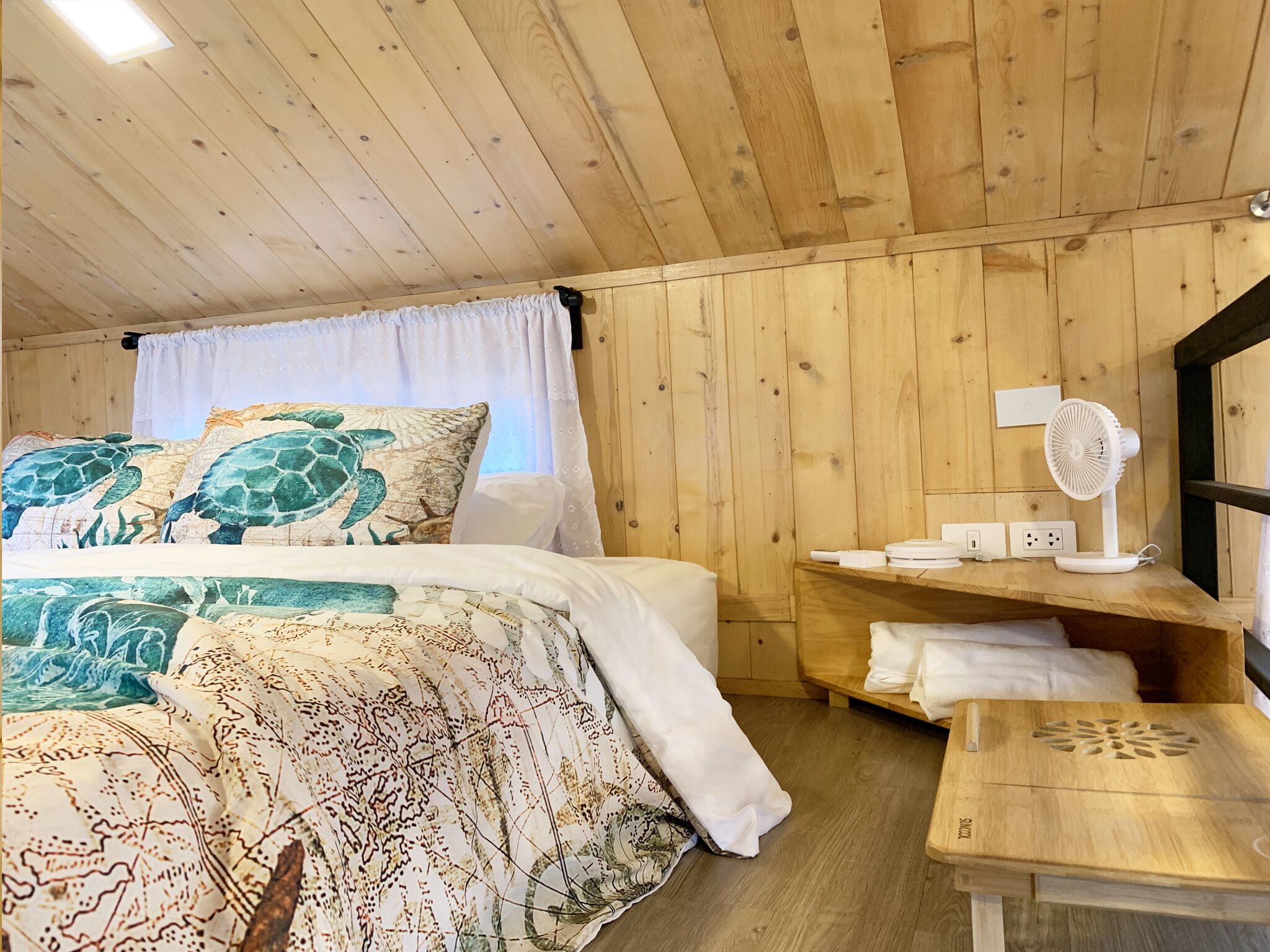 A wooden cabin room with a bed, a small table, a fan, and a window with curtains.