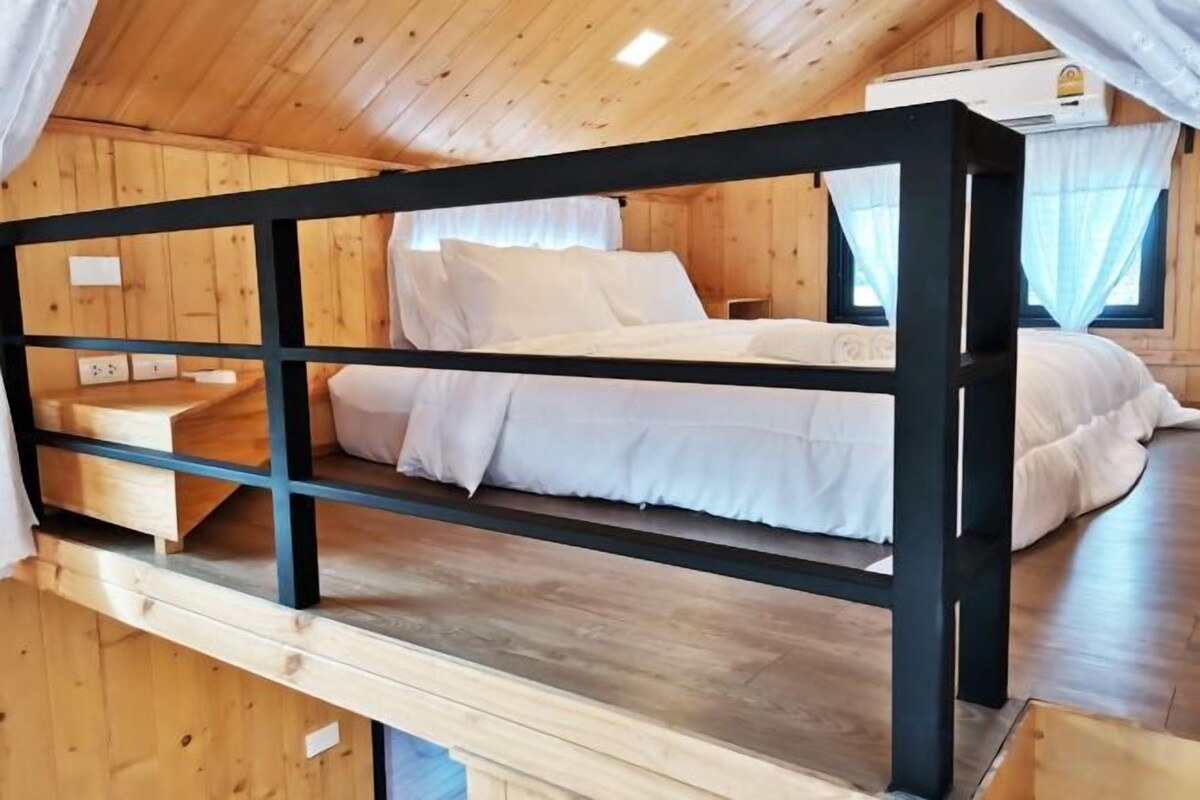 Una cama litera con estructura de madera y ropa de cama blanca, una pequeña mesa de madera y un ventanal con cortinas.