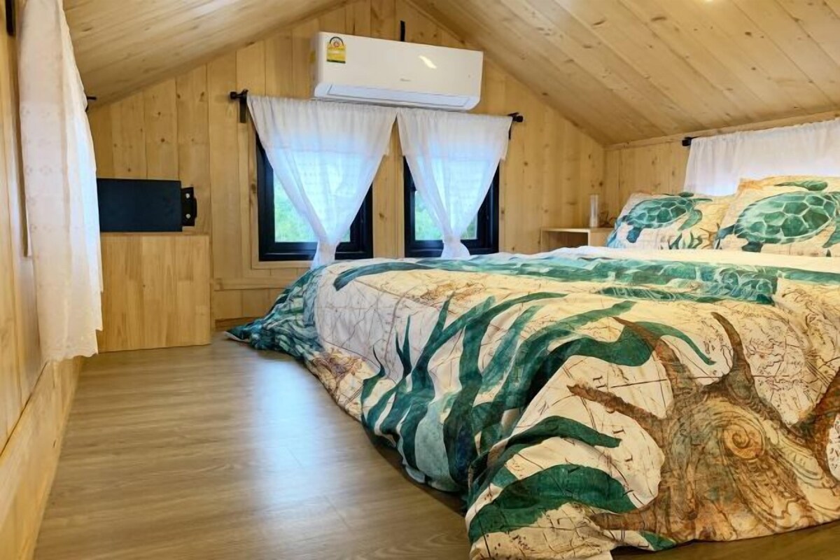 Una habitación de madera con una cama, dos ventanas con cortinas e un equipo de aire acondicionado.