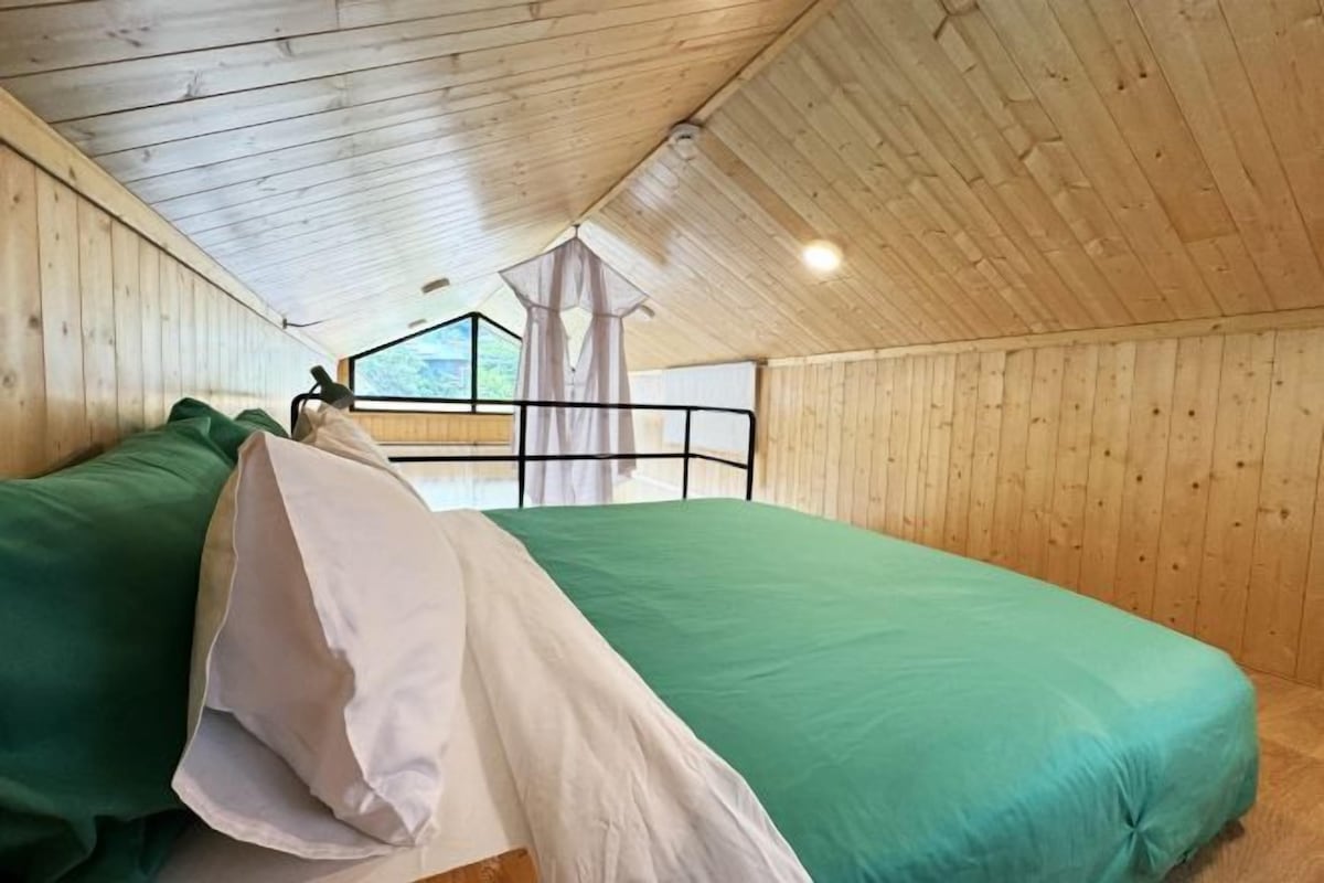 Una habitación con techo y paredes de madera, una cama con ropa de cama verde y blanca, y una ventana que ofrece vistas a la vegetación.