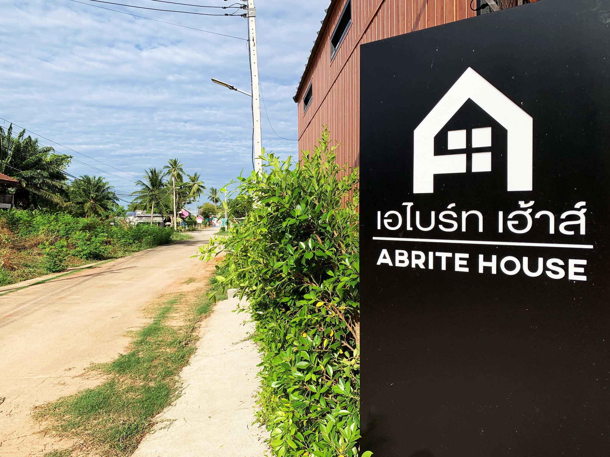 Un panneau indiquant « Abrite House », avec un symbole représentant une maison, placé au bord d’un chemin de terre, avec de la végétation et un bâtiment en arrière-plan.