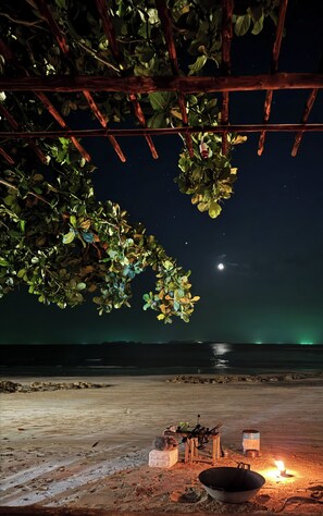 Escena nocturna con una pérgola de madera, una fogata y vistas al océano bajo un cielo estrellado.