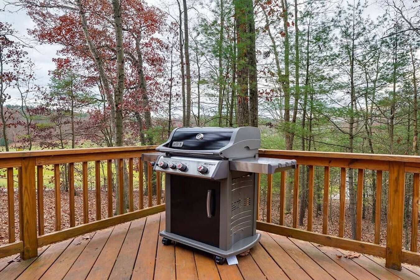 Ein tragbarer Grill auf einer Holzterrasse mit Geländer, umgeben von Bäumen.