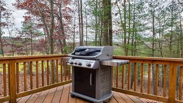 Ein tragbarer Grill auf einer Holzterrasse mit Geländer, umgeben von Bäumen.