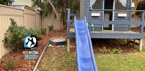 Un jardin avec un toboggan, une zone délimitée par une clôture et une maison dotée d’un balcon.