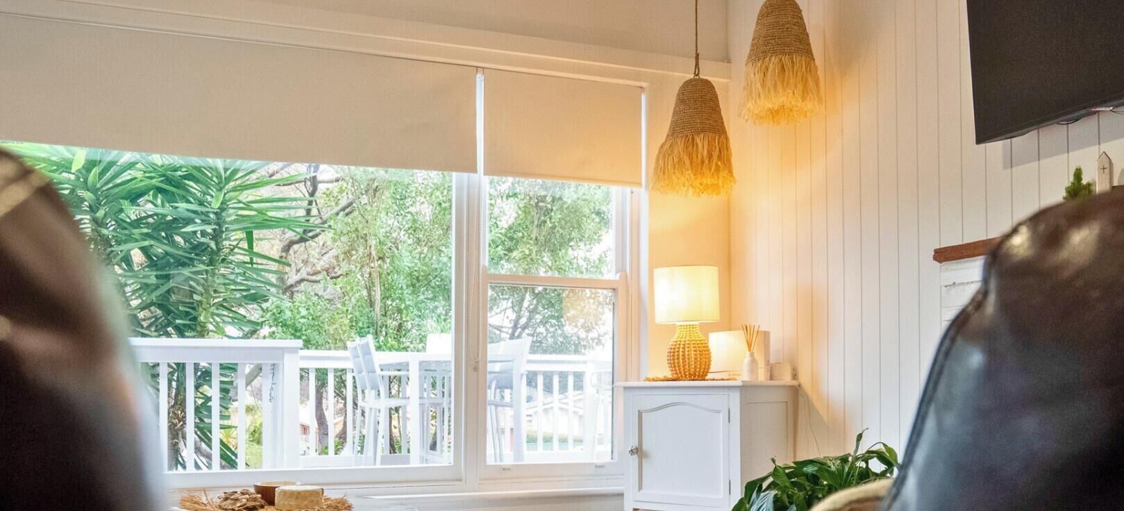 A living room with a leather sofa, a small table, and a lamp. There is a view of a white fence and greenery outside.