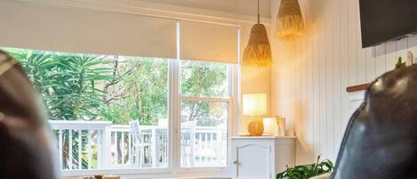 A living room with a leather sofa, a small table, and a lamp. There is a view of a white fence and greenery outside.