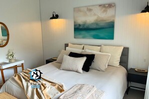 A bedroom with a large bed, two bedside tables, a nightstand, a mirror, and a wall painting. - Saltwater + Sunsets – Seaside Haven with Ocean Views and Family-Friendly Charm (Victor Harbor)