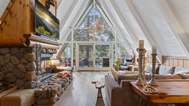 A cozy loft-style room with a stone fireplace, a large TV, and a view of the outdoors. - Rocky Point Lakefront | Private Dock & Hot Tub (Lake Arrowhead)