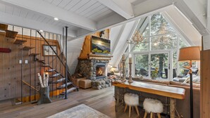 A cozy interior with a stone fireplace, wooden staircase, and a large window offering a view of trees. - Rocky Point Lakefront | Private Dock & Hot Tub (Lake Arrowhead)