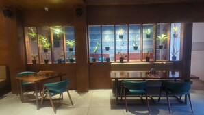 A restaurant with a wooden interior, teal chairs, and a glass partition with potted plants. - The Pramukh Hotel (Sinor)
