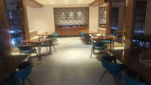A dining area with multiple tables and chairs, a large decorative wall, and a view of the outside through the windows. - The Pramukh Hotel (Sinor)