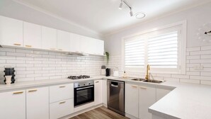 Une cuisine moderne avec des meubles blancs, une cuisinière à gaz, un lave-vaisselle et une fenêtre avec des stores.