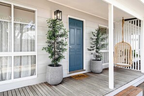 Varanda com porta azul, plantas em vasos, cadeira suspensa e tapete de boas-vindas.