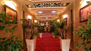 Pasillo de hotel con alfombra roja, plantas en macetas, obras de arte enmarcadas y un techo decorativo.