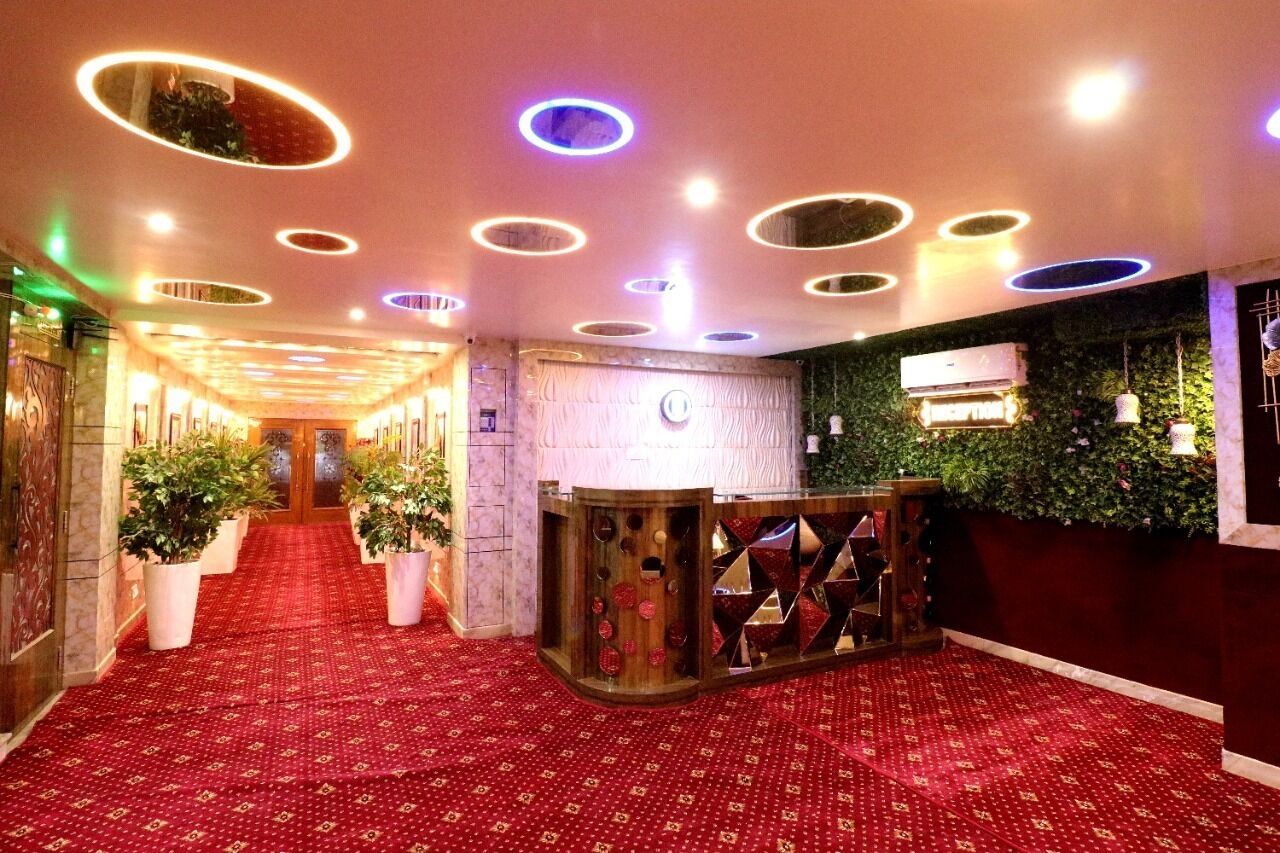Un hall d’hôtel avec un tapis rouge, des luminaires circulaires au plafond, un comptoir de réception et des plantes en pot.