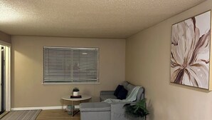 A living room with a grey sectional sofa, a small round table with a plant, a window with blinds, and a large abstract painting on the wall.