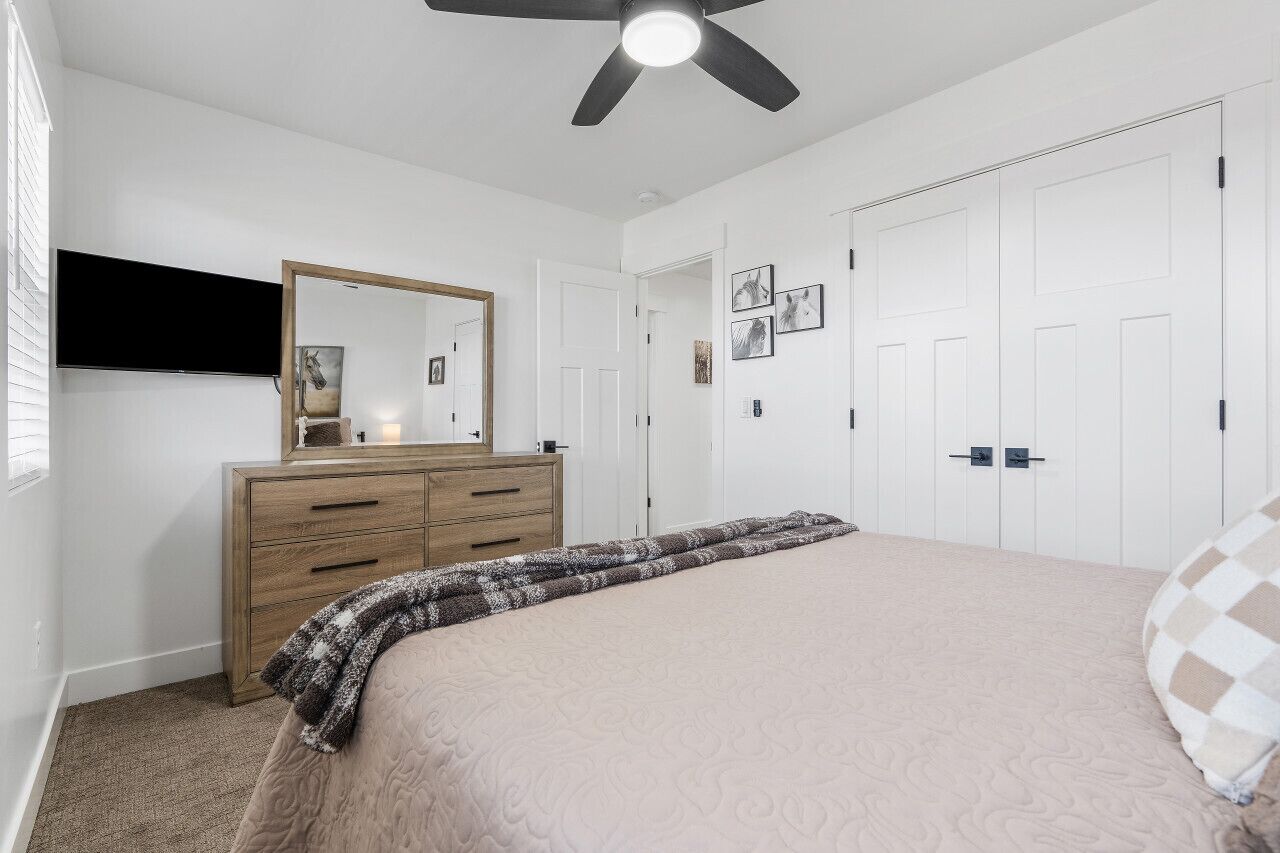 Un dormitorio con una cama, un aparador, un ventilador de techo, un televisor montado en la pared y una puerta con espejo.