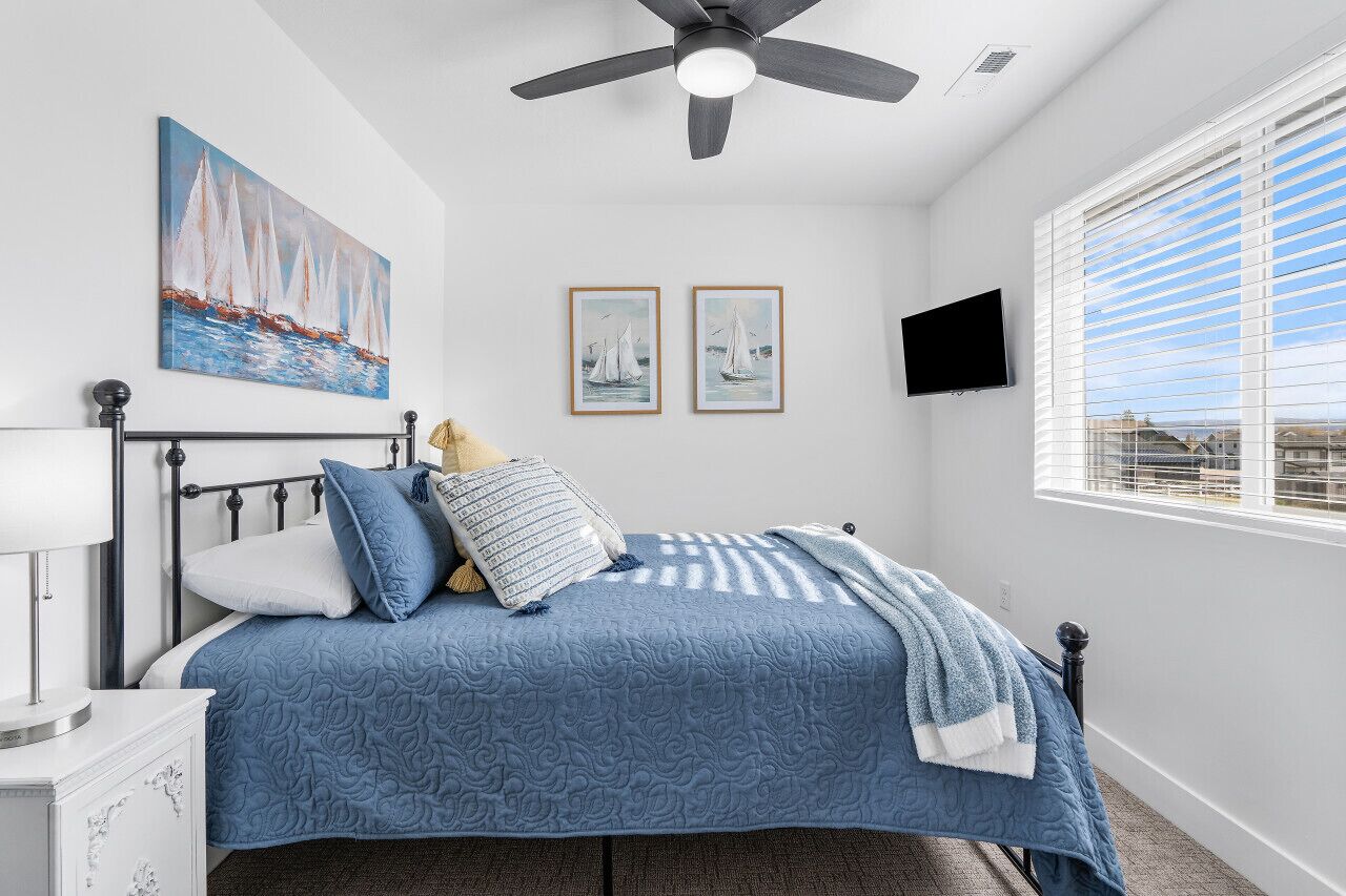 Une chambre à coucher avec un lit, un ventilateur de plafond, une fenêtre avec des stores et des œuvres d’art accrochées aux murs.