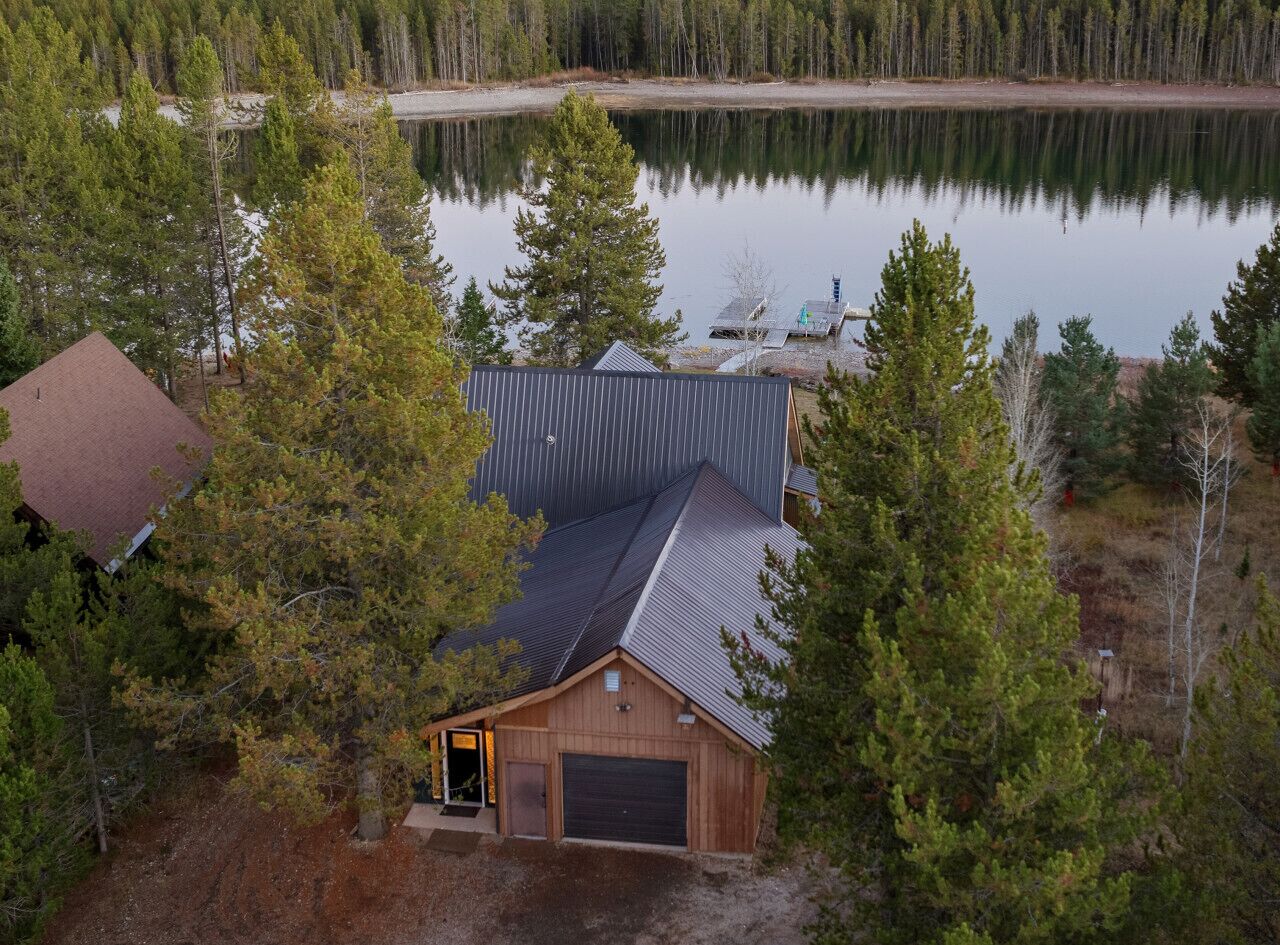 Une cabane avec un toit en métal, un garage et un quai s’avançant dans un lac paisible, le tout entouré d’une forêt dense.