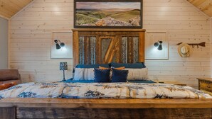 Een slaapkamer met een houten plafond, een bed met beddengoed met een patroon, een aan de muur bevestigd schilderij van een landschap, en twee wandlampen.