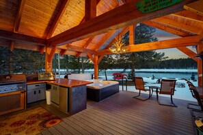 Une terrasse en bois avec un espace cuisine, comprenant un barbecue et un jacuzzi, donnant sur un paysage enneigé.