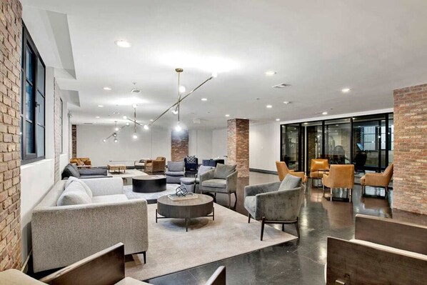 A modern lounge area with sofas, armchairs, and a coffee table, featuring exposed brick walls and large windows.