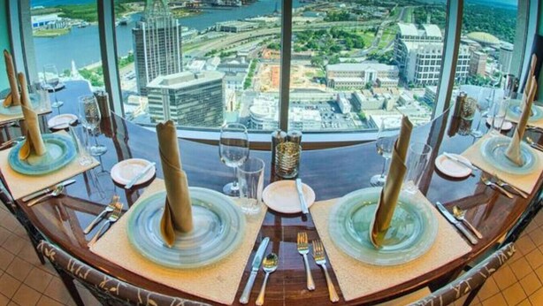 Une table à manger, dressée avec des assiettes, des couverts et des verres, se détache sur un arrière-plan composé d’une vue sur la ville et sur l’eau.