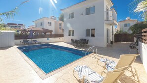 A swimming pool with lounge chairs and a table, surrounded by a white building with balconies. - Protaras Blue River Villa Br26 (Περνέρα)