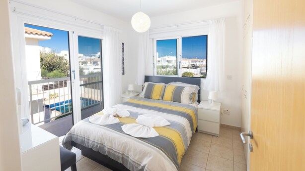 A bedroom with a bed, bedside tables, a view of the city, and a balcony with a pool. - Protaras Blue River Villa Br26 (Περνέρα)