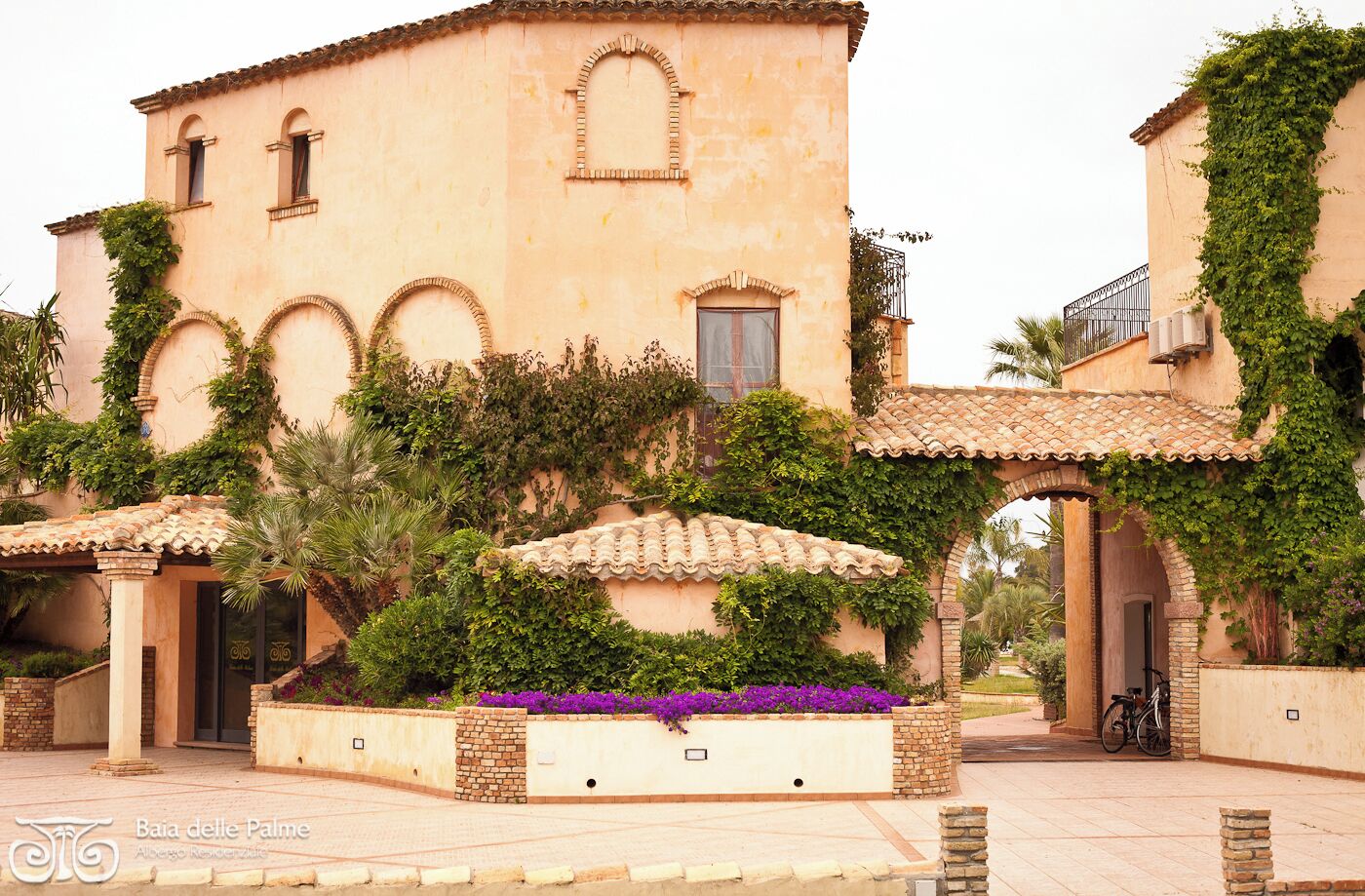 A building with arched windows, a tiled roof, and a garden with purple flowers.