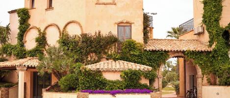 A building with arched windows, a tiled roof, and a garden with purple flowers.