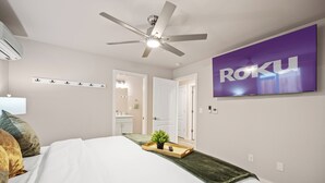 Een slaapkamer met een bed, een plafondventilator, een televisie waarop 'ROKU' staat en een zichtbare badkamer door een geopende deur.