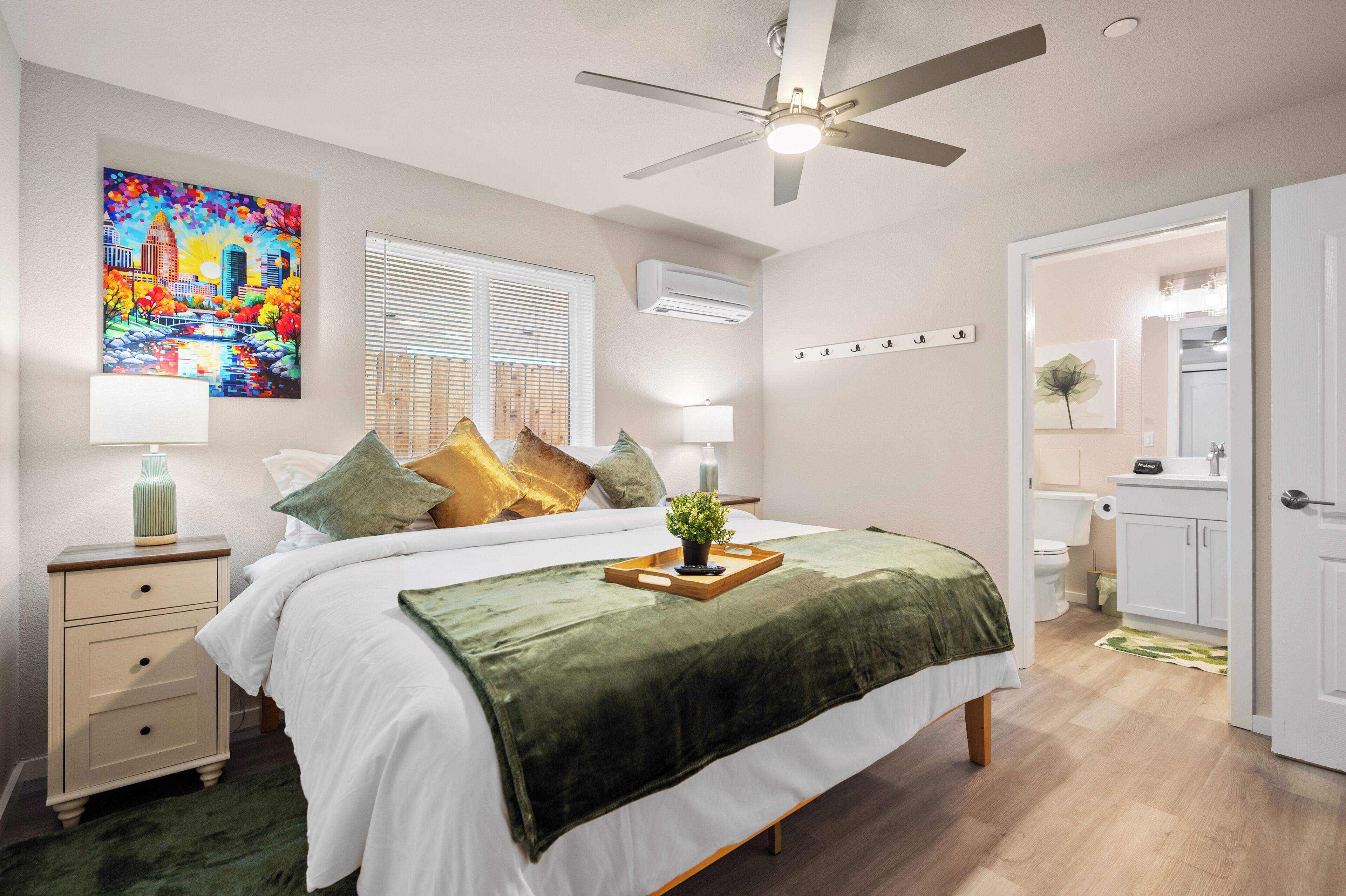 A bedroom with a bed, bedside tables, a ceiling fan, a window with blinds, an air conditioning unit, a bathroom with a toilet, and a door leading to another room.