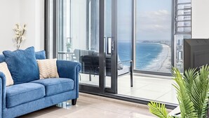 A modern living room with a blue sofa, a glass door leading to a balcony with a view of the ocean and city skyline. - Oceanfront Escape! 2 Beds Home w/ Gym  & Carpark (Surfers Paradise)