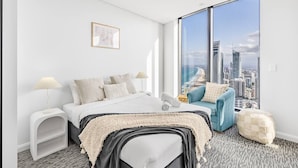 A modern bedroom with a large bed, a blue armchair, a white side table, and a view of a city skyline through large windows. - Oceanfront Escape! 2 Beds Home w/ Gym  & Carpark (Surfers Paradise)