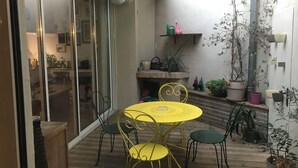 En terrasse med et gult bord og stole, omgivet af krukker med planter og en skydedør til et indendørs område.