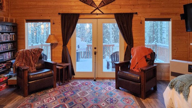 A cozy cabin interior with a wooden ceiling, two leather chairs, a patterned rug, a bookshelf, and a large window with curtains.
