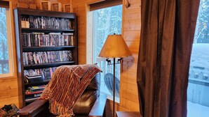 A cozy cabin interior with a leather chair, a bookshelf, a lamp, and a large window with curtains.