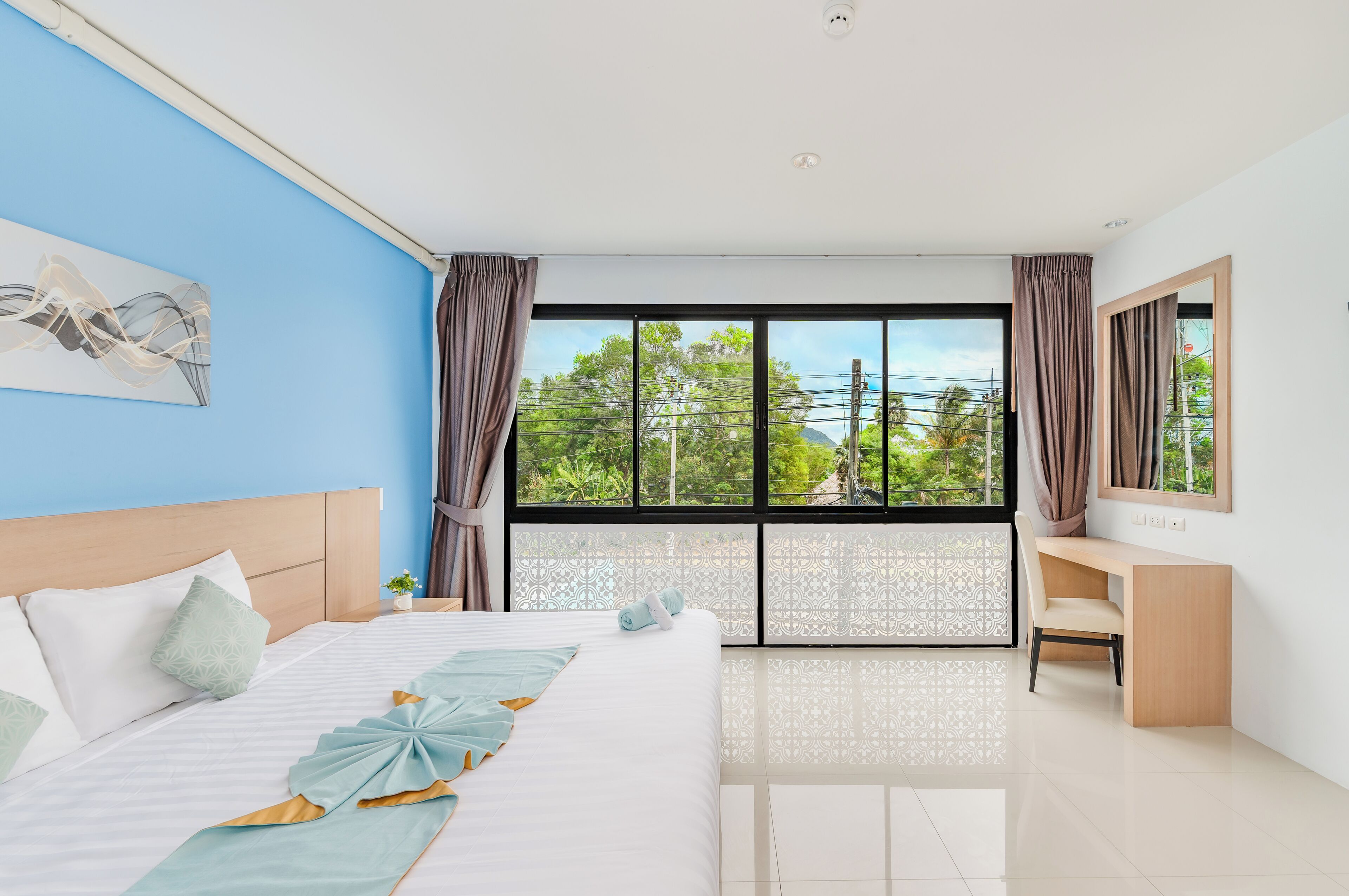 A modern hotel room with a large bed, a desk, and a view of greenery outside.