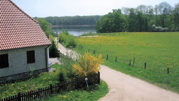 A rural house with a red-tiled roof, a dirt path leading to a field, and a body of water in the background. - Apartment 'Ferienwohnung Stadermann' with Private Terrace, Private Garden and Balcony (Walow-Strietfeld)