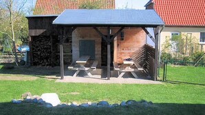 A covered outdoor seating area with picnic tables, a stone wall, and a red-roofed building in the background. - Apartment 'Ferienwohnung Stadermann' with Private Terrace, Private Garden and Balcony (Walow-Strietfeld)