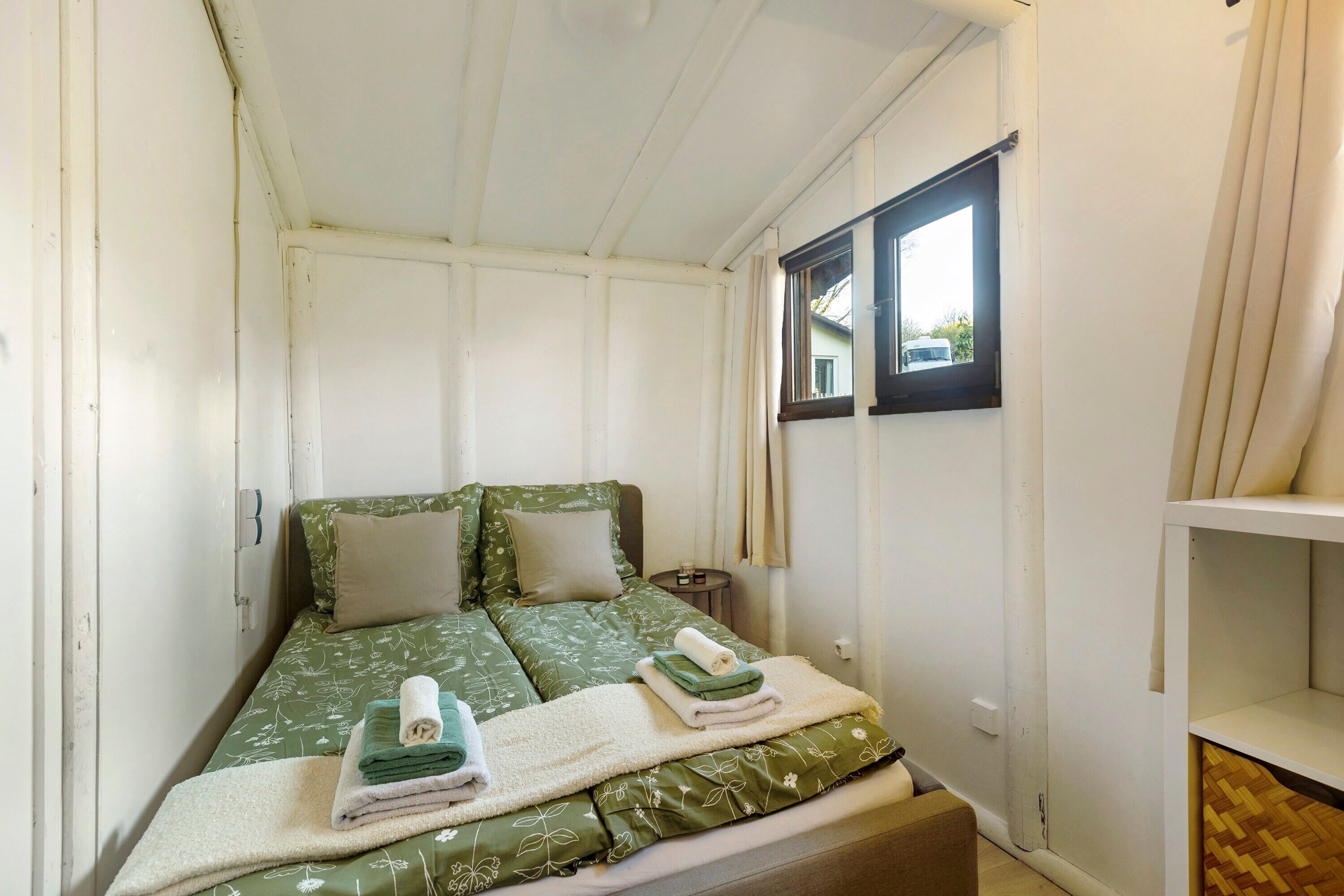 A small, single-bed room with a green floral bedspread, a white pillow, and a neatly made bed with folded towels.