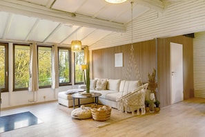 A cozy living room with a sofa, a coffee table, a hanging light fixture, and a view of trees through large windows.