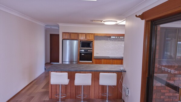 A kitchen with wooden cabinets, a stainless steel refrigerator, an oven, and a microwave. There are three white bar stools at the counter. - Gateway to Shoalhaven. Winter specials available now. (Bomaderry)