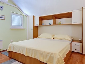 A bedroom with a bed, a nightstand, a shelf with items, and a window with blinds.