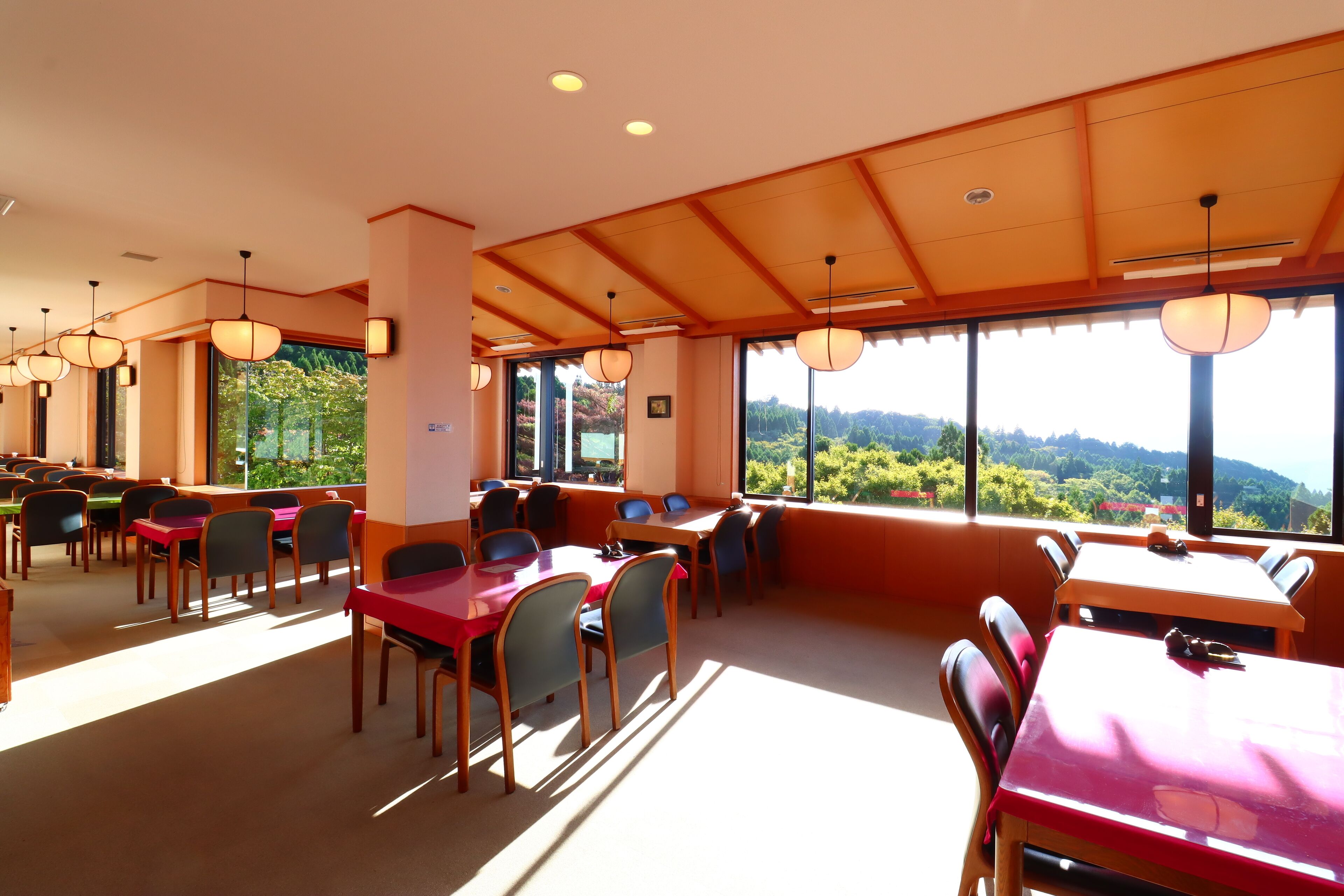 A dining area with tables and chairs, large windows with a view of greenery, and pendant lights.