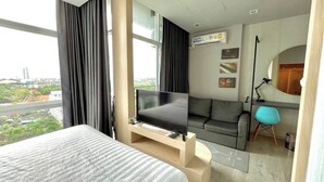 Quarto de hotel moderno com uma cama grande, uma TV sobre um suporte de madeira, um sofá, uma escrivaninha com cadeira e vista para a paisagem urbana.